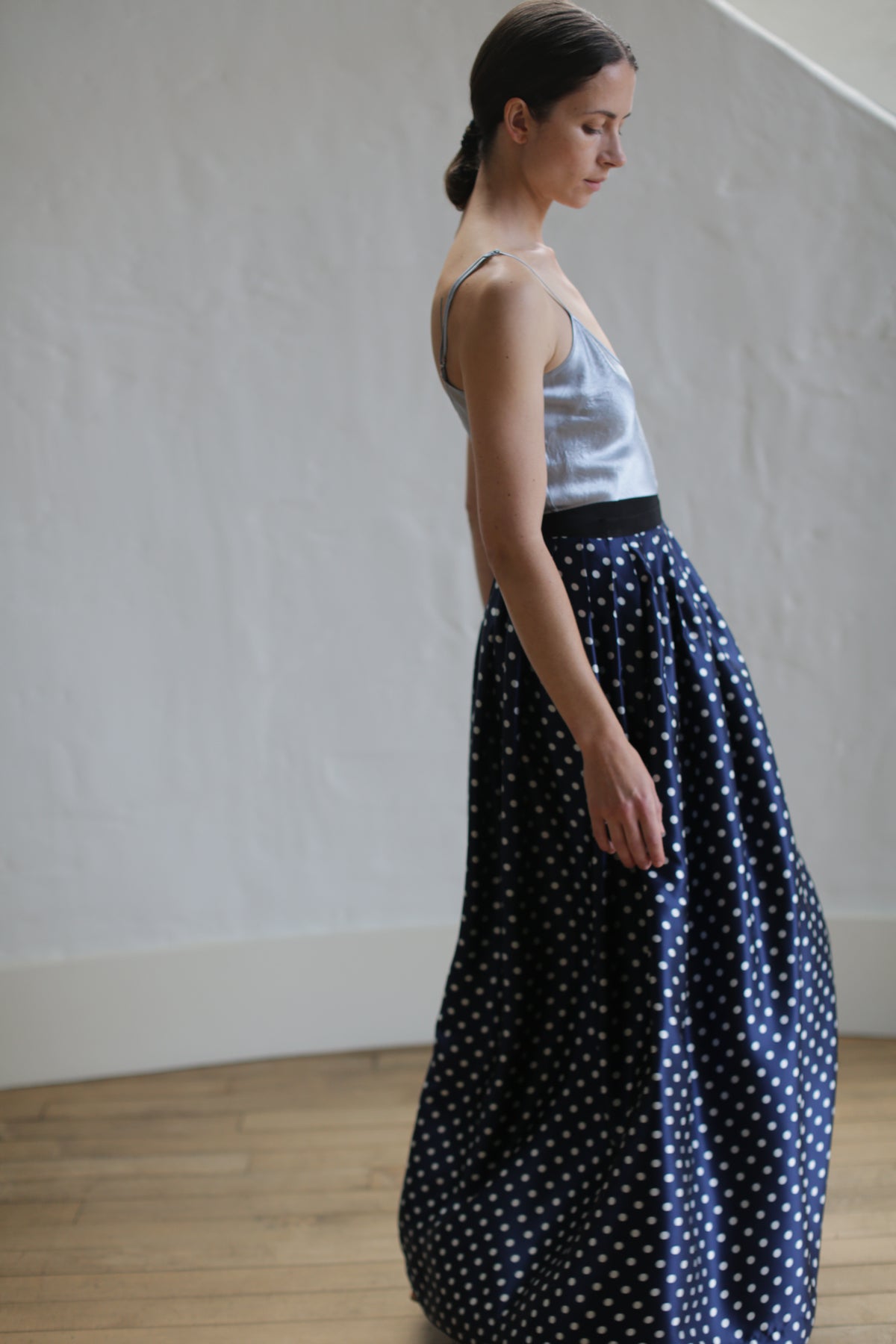 A woman with her hair tied back is standing and looking to the side. She wears a silver camisole and a Charmeuse Pleated Long Wrap Skirt | Navy Polkadot. She is in a minimalist room with white walls and wooden flooring.