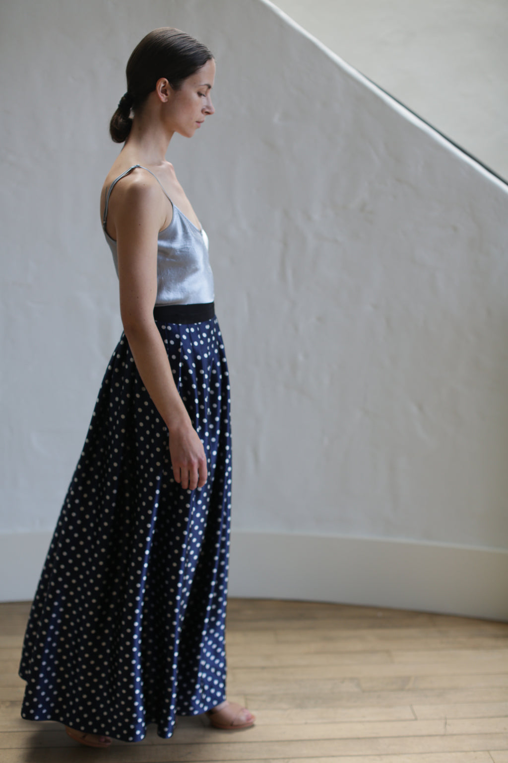 A woman with her hair tied back stands in profile, wearing a grey camisole top and a Charmeuse Pleated Long Wrap Skirt | Navy Polkadot. She is barefoot on a wooden floor, and there is a white wall with a slight curve in the background.