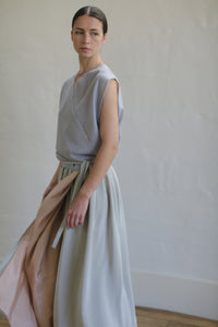 A woman wearing a sleeveless gray top and a long, flowing Silk Reversible Pleated Long Wrap Skirt | Celadon/Blush stands against a neutral background. She has her hair pulled back and is looking to the side, with a relaxed yet thoughtful expression.