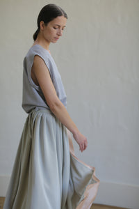 A person with long hair tied back is standing and looking down. They are wearing a sleeveless top and a flowing, Silk Reversible Pleated Long Wrap Skirt | Celadon/Blush. The background is plain and light-colored, adding a minimalist and serene feel to the image, emphasizing its zero-waste design approach.