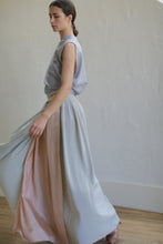 Load image into Gallery viewer, A person with dark hair tied back in a low ponytail is wearing a flowing, sleeveless gray top and a Silk Reversible Pleated Long Wrap Skirt | Celadon/Blush. The 100% silk fabric moves gracefully as they walk on a light wood floor against a plain white wall, with one arm slightly raised.