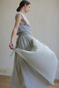 A woman in a sleeveless gray top and a Silk Reversible Pleated Long Wrap Skirt | Celadon/Blush made from 100% silk stands in a minimalist setting with hardwood flooring and an off-white wall. She holds part of her skirt, creating a sense of motion and elegance. Her hair is pulled back in a low bun.
