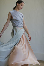 Load image into Gallery viewer, A person with a serious expression is posing, standing with one arm lowered and the other slightly raised. They are wearing a sleeveless, asymmetrical gray top and a Silk Reversible Pleated Long Wrap Skirt in Celadon/Blush. The design features zero-waste principles, and the background is plain and neutral.