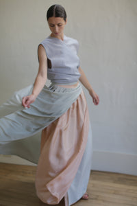 A woman with her hair tied back twirls gracefully, wearing a sleeveless top and a Silk Reversible Pleated Long Wrap Skirt | Celadon/Blush made from 100% silk with layers of light blue and peach fabric. She stands on a wooden floor against a plain, light-colored wall, embodying an elegant zero-waste design ethos.