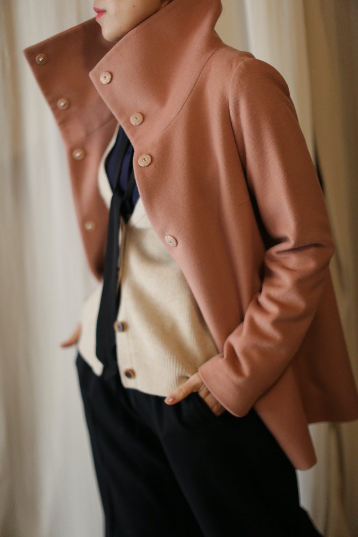 A person stands with hands in pockets, attired in a beige shirt with a black tie, and wearing the Stamford II light brown jacket, distinguished by its popped high collar and button details. They pair this ensemble with dark pants. The individual is slightly turned to the side, facing left against a softly blurred, neutral-toned background.