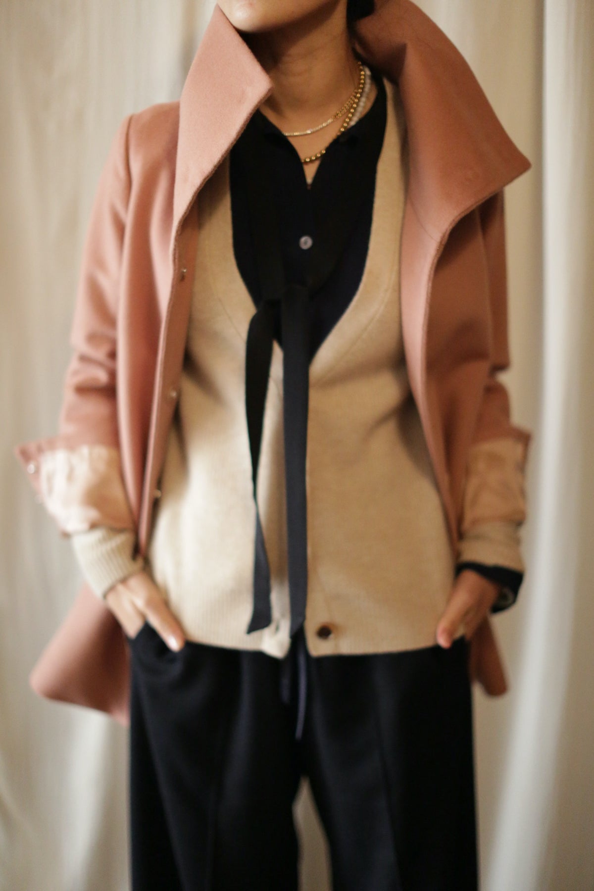 A person is dressed in a stylish outfit featuring a beige cardigan layered over a black button-up shirt with a bow tie, black pants, and the rose-colored Stamford II jacket with its high collar turned up. They are standing with their hands in their pockets, facing slightly to the left.