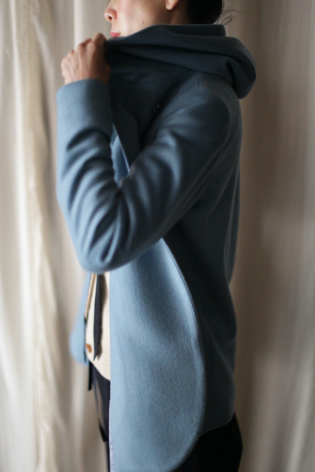 Wearing the Mitchley, a person in a light blue double-breasted jacket lifts the collar near their face and stands sideways against a softly lit, light-colored curtain backdrop.