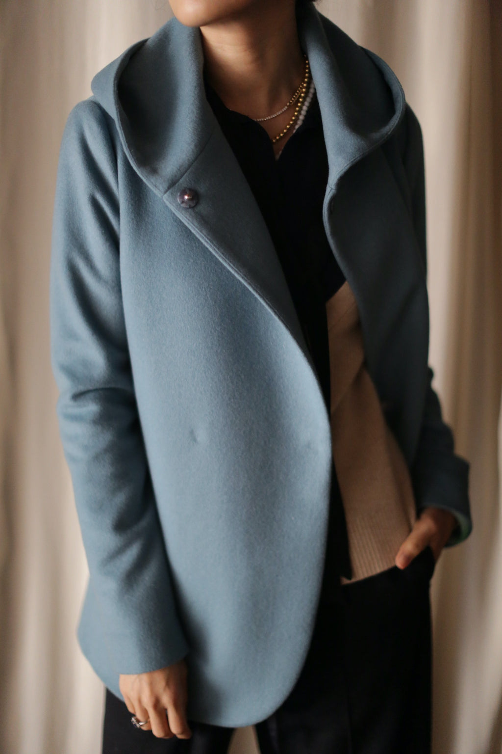 Wearing the Mitchley light blue double-breasted jacket with a wide collar over a beige sweater and black top, accessorized by gold necklaces, this look exudes luxurious everyday style against a softly blurred background.