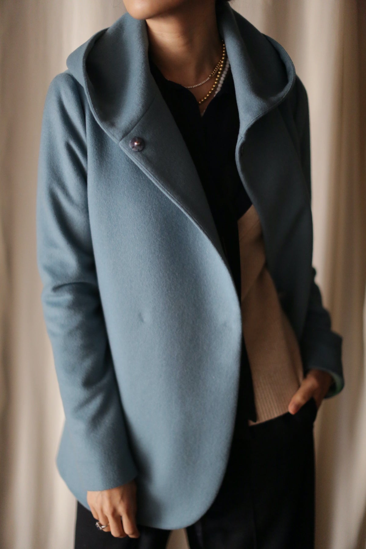 Wearing the Mitchley light blue double-breasted jacket with a wide collar over a beige sweater and black top, accessorized by gold necklaces, this look exudes luxurious everyday style against a softly blurred background.