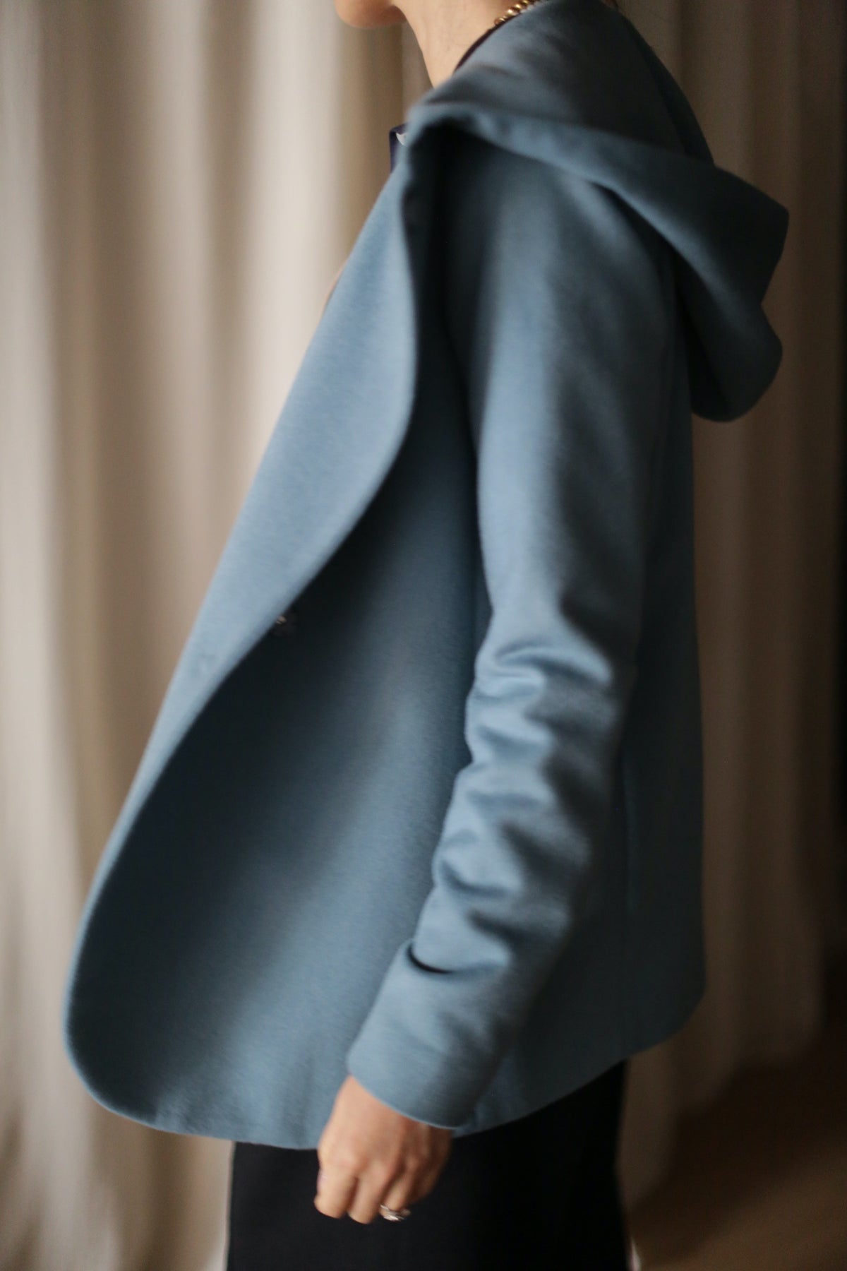 Someone stands sideways in the Mitchley jacket, a luxurious everyday piece with a light blue cashmere hood. Their left arm is slightly bent and hand visible. The softly blurred background features light-colored curtains.