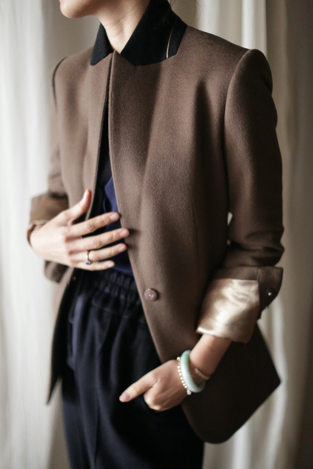A person wearing the Sloane blazer with structured shoulders and cuffed sleeves over a black shirt and dark pants stands in front of a light-colored backdrop. One hand is in their pocket, and the other touches the front of the blazer, showing a ring and bracelets.