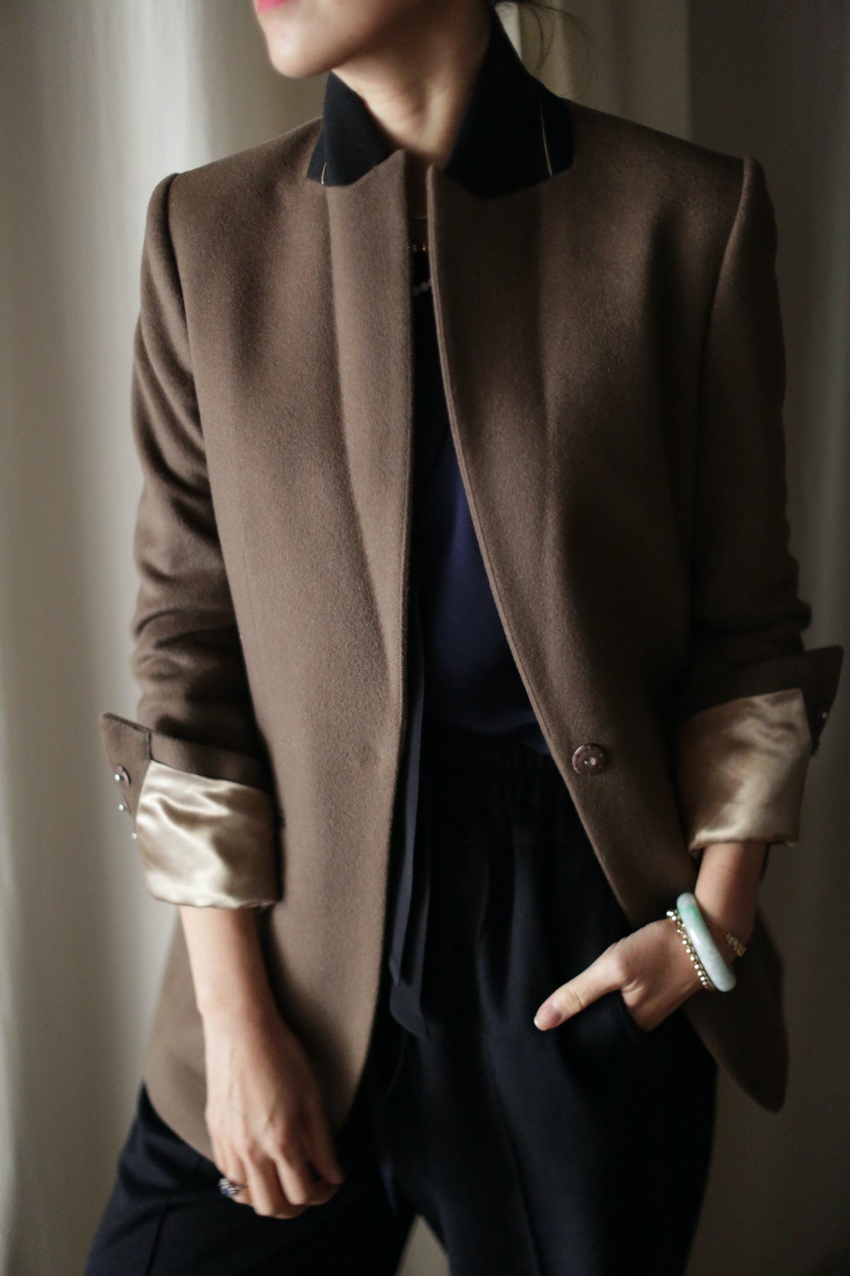 Sloane is showcased in a dark brown coat featuring structured shoulders, with the sleeves stylishly rolled up to reveal a light-colored lining. Paired with dark pants and a bracelet adorning the left wrist, the ensemble exudes sophistication. The individual strikes a casual pose, standing with one hand in their pocket, though their face remains out of view.