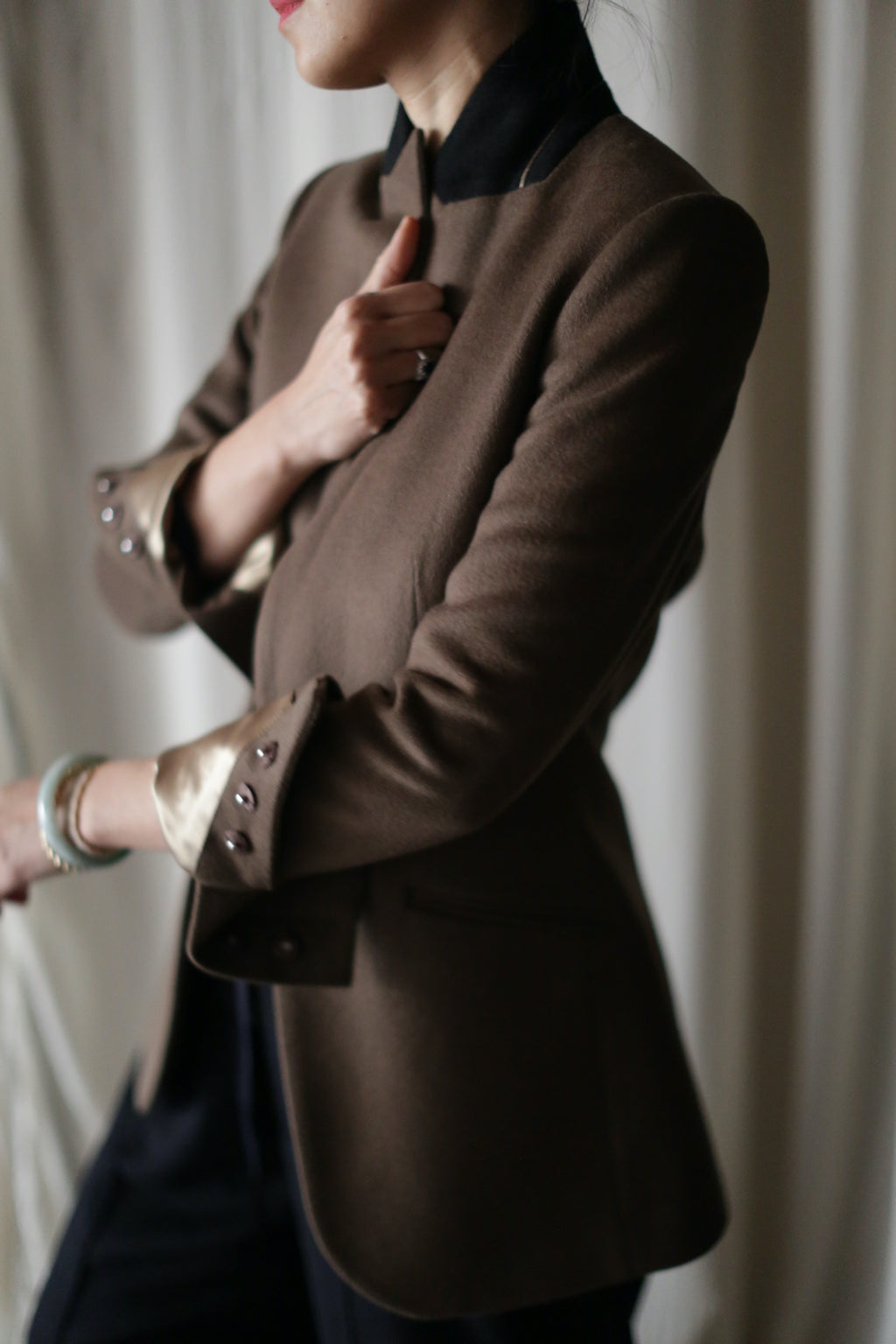 A person wearing the "Sloane" blazer, characterized by its brown hue, rolled-up sleeves, and structured shoulders, paired with a black shirt underneath, is holding the lapel of the blazer. The image is taken from the side, focusing on the upper body against a softly blurred, neutral backdrop.