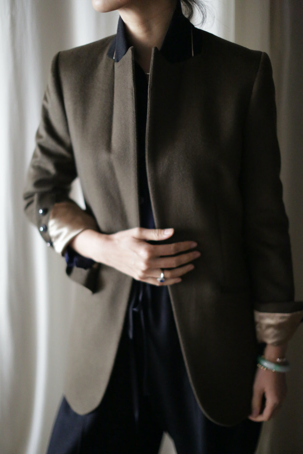 A person is dressed in the "Sloane" blazer, featuring a dark green color with structured shoulders and black and beige accents. Their left hand, adorned with a ring and bracelet, rests on their midsection. The light-colored curtain serves as the background, while the person's face remains unseen in the image.