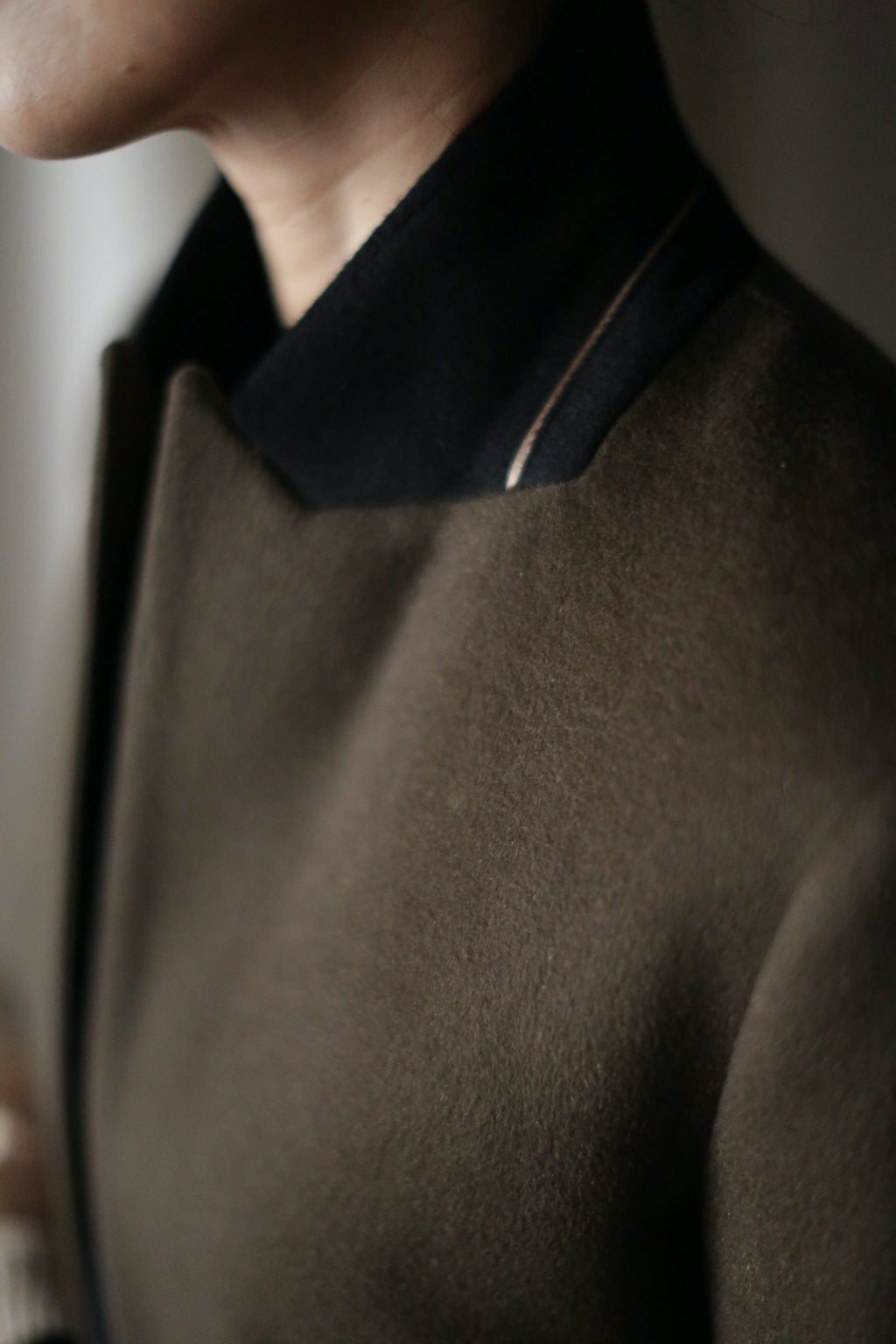 Close-up of a person wearing the Sloane coat in dark green or olive, accented by a black collar. The image captures only the lower part of the face and the top portion of this equestrian-length coat, highlighting the rich textures and colors of its woven cashmere fabric. The background is blurred.
