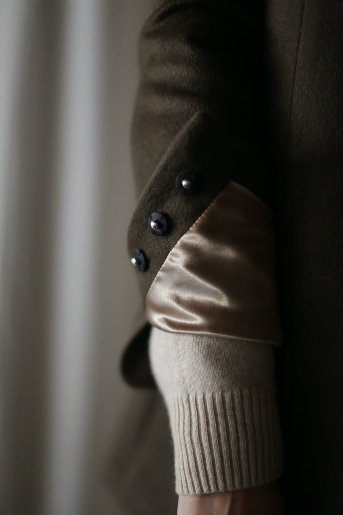 Close-up of a person's arm showing detailed fashion elements. The person is wearing the Sloane, a beige sweater underneath a dark brown coat with structured shoulders. The coat boasts turned-up sleeves revealing a shiny beige lining and three black buttons at the cuff.