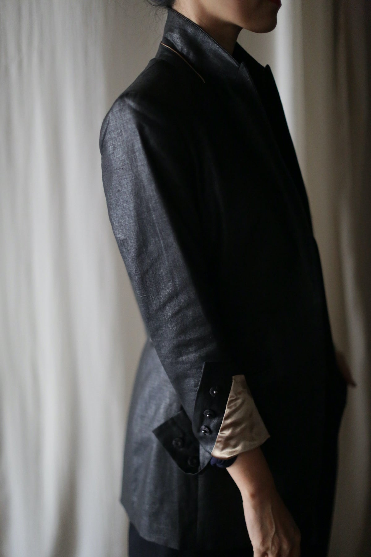 A person is wearing a dark-colored Waxed Linen Sloane tuxedo jacket with the sleeves rolled up to reveal a beige lining. The shot captures the person from neck to mid-thigh, with their head slightly turned away from the camera. The background is softly lit, featuring light-colored curtains.