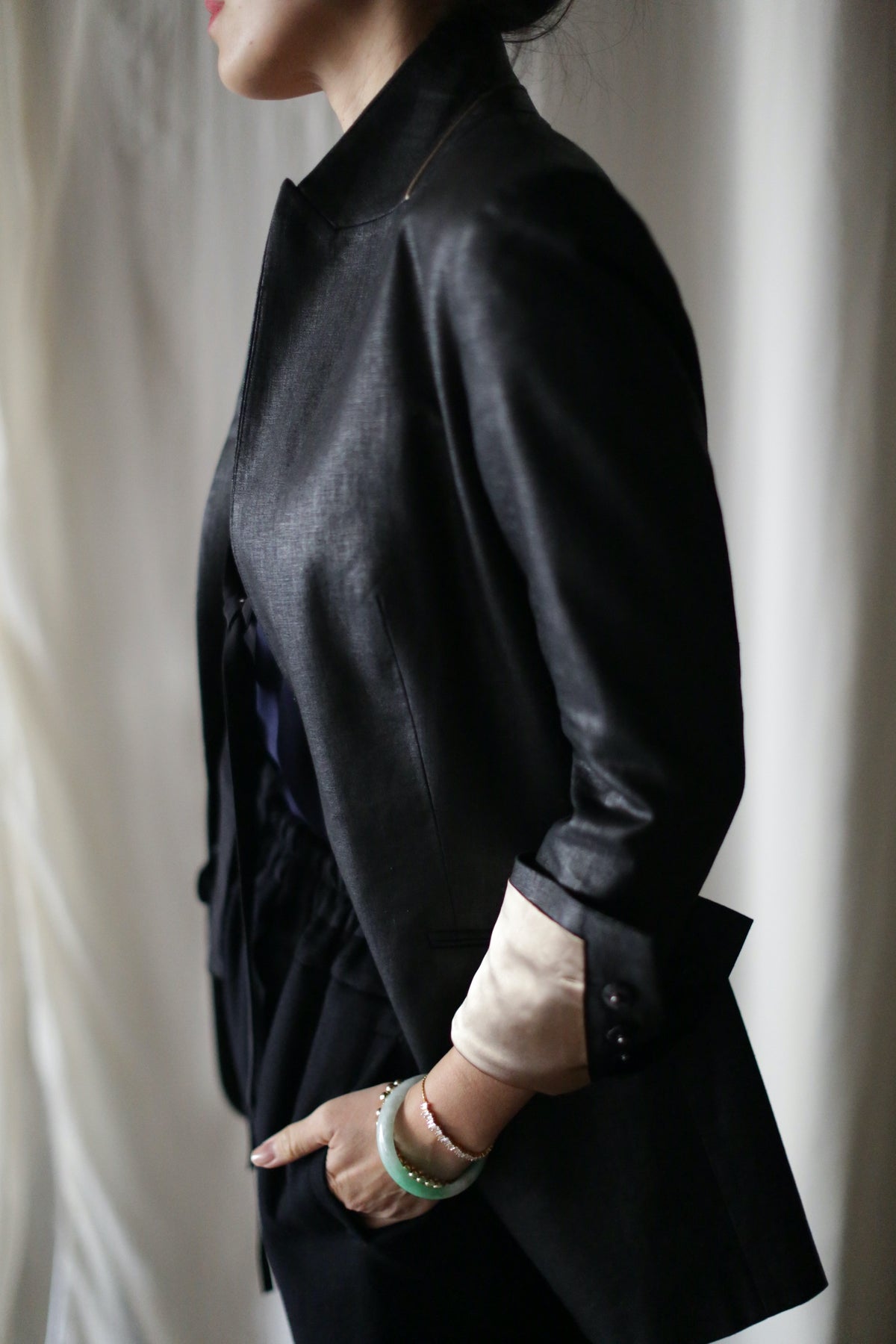 A person wearing a Waxed Linen Sloane tuxedo jacket with rolled-up sleeves, revealing a light-colored inner lining. They stand with one hand in their pocket, adorned with bracelets on their wrist. The background is plain and light-colored, highlighting the outfit's details and the breathable wax finish.
