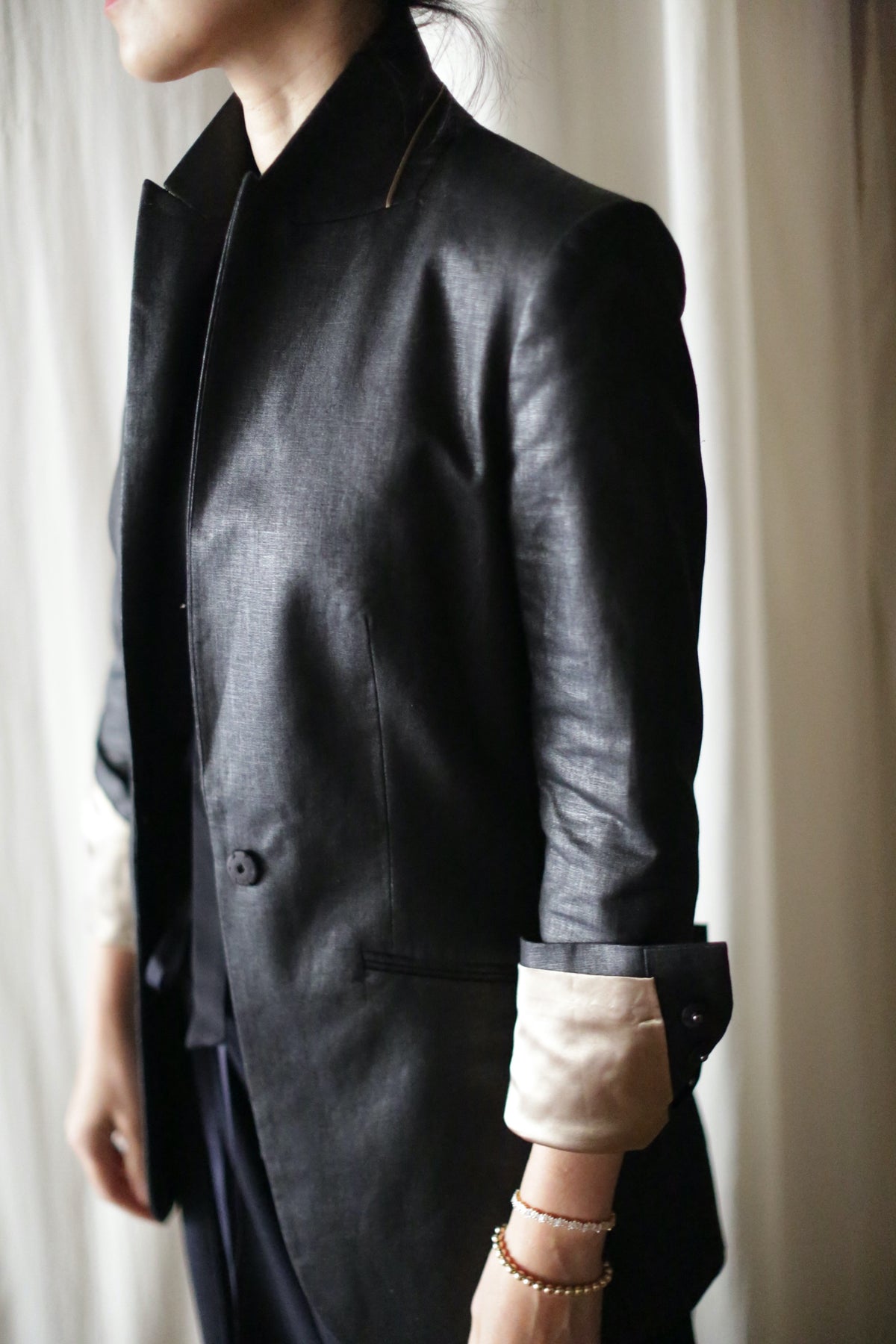 A person wearing the Waxed Linen Sloane blazer with the sleeves rolled up, revealing a light-colored inner lining. The individual, whose head is turned away, stands against a light-colored curtain backdrop, sporting subtle wrist jewelry to complete the outfit.