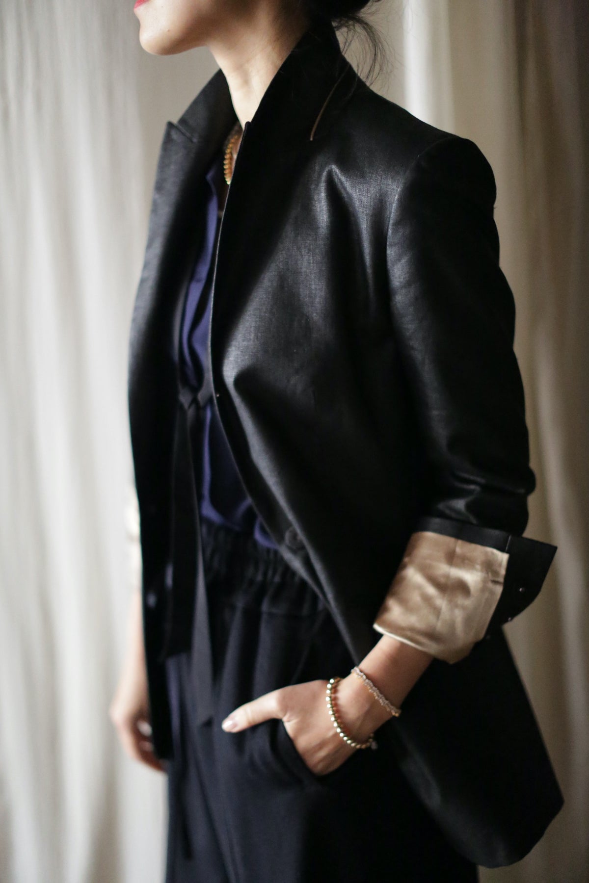 A person dressed in a Waxed Linen Sloane tuxedo jacket with rolled-up sleeves and navy blue clothing underneath stands with one hand in their pocket. Their head is slightly turned away from the camera, and the background features a light, draped fabric.