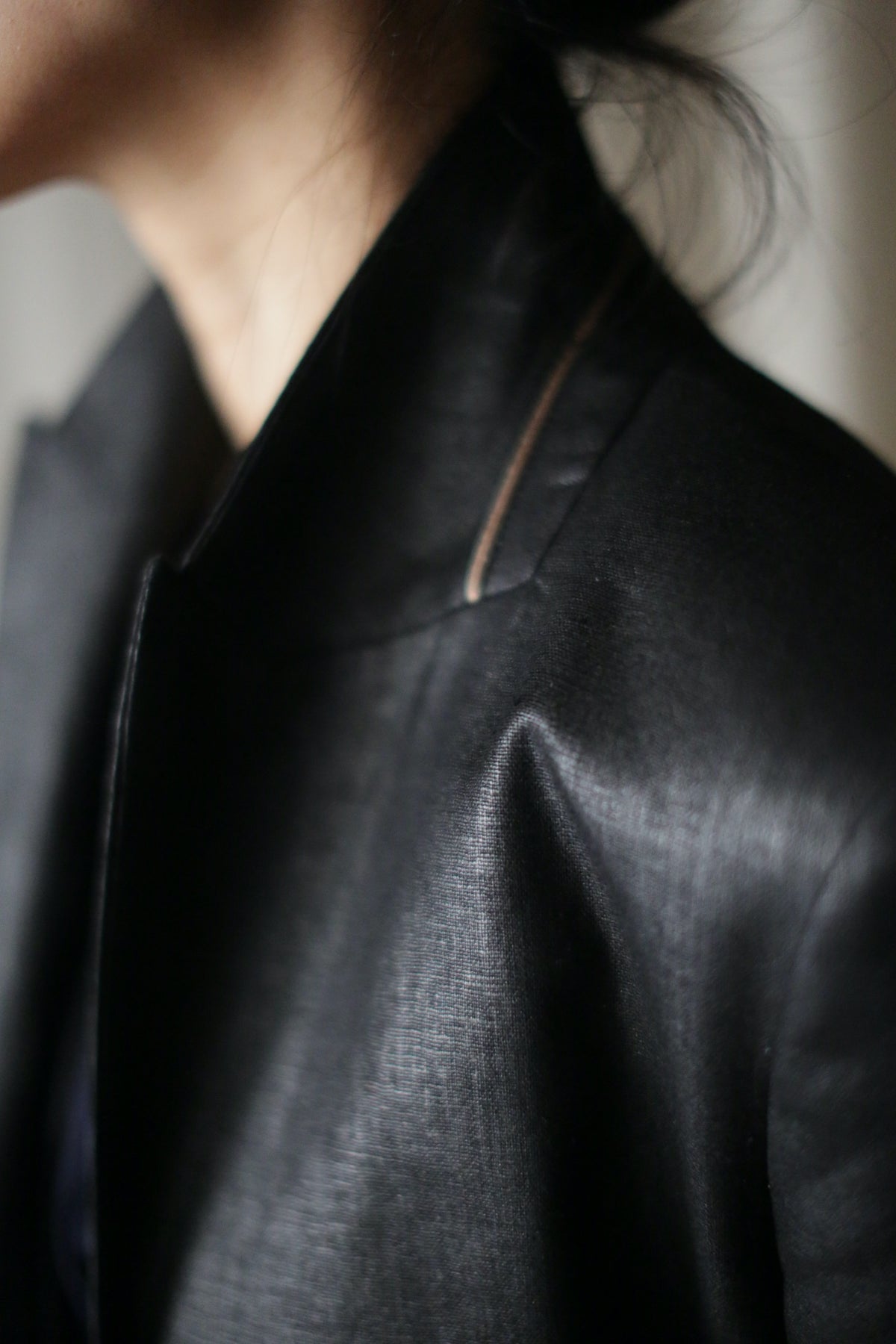 Close-up of a person wearing the Waxed Linen Sloane jacket in black, featuring a high collar. The focus is on the shoulder and upper chest area, highlighting the fabric's sheen and texture. The background is out-of-focus, with the person's face partially out of the frame.