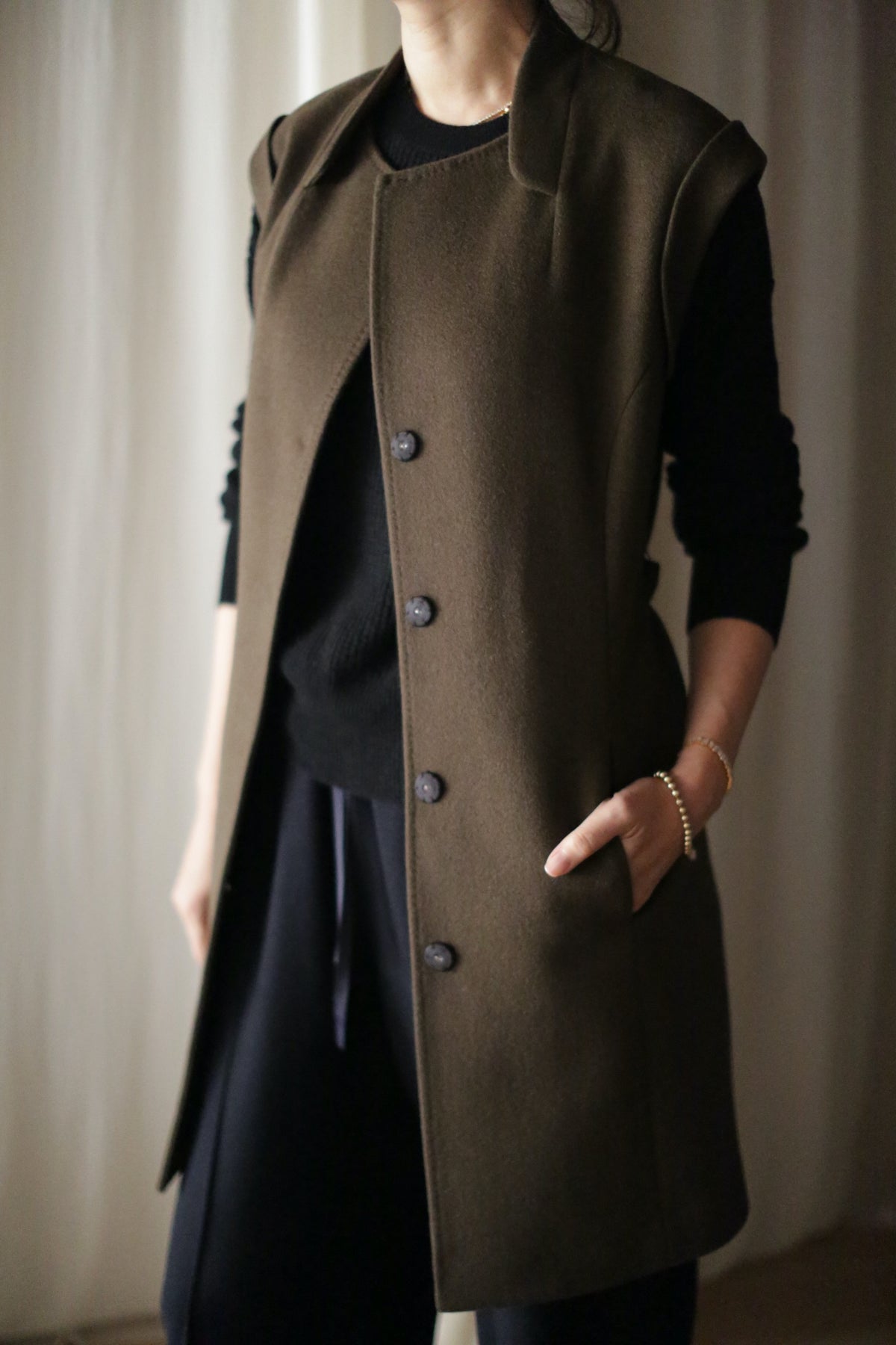 A person wears the Lexington, a long sleeveless dark olive vest-coat, over a black top and pants, standing with one hand in a pocket against a softly blurred background.