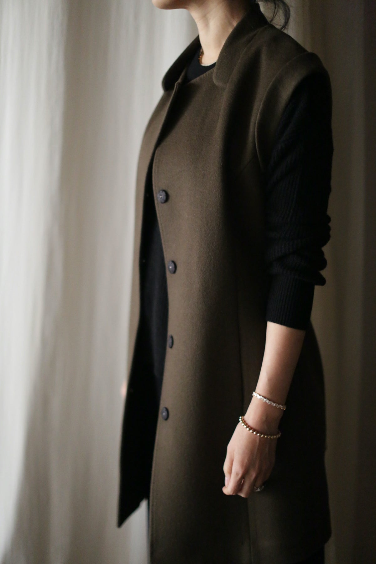 A person stands sideways, partly out of frame, wearing the Lexington long olive-green cashmere vest coat over a black top. They accessorize with bracelets on their right wrist and stand before a softly lit, neutral curtain.