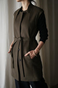 Indoors, a person wears the elegant Lexington cashmere vest coat featuring a belted waist over a black long-sleeve top, hands in pockets. A softly blurred background enhances the coat’s refined appeal.