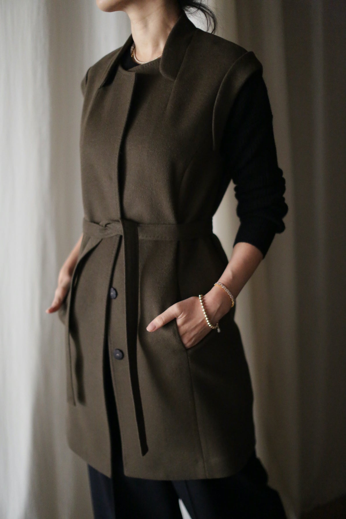 Indoors, a person wears the elegant Lexington cashmere vest coat featuring a belted waist over a black long-sleeve top, hands in pockets. A softly blurred background enhances the coat’s refined appeal.