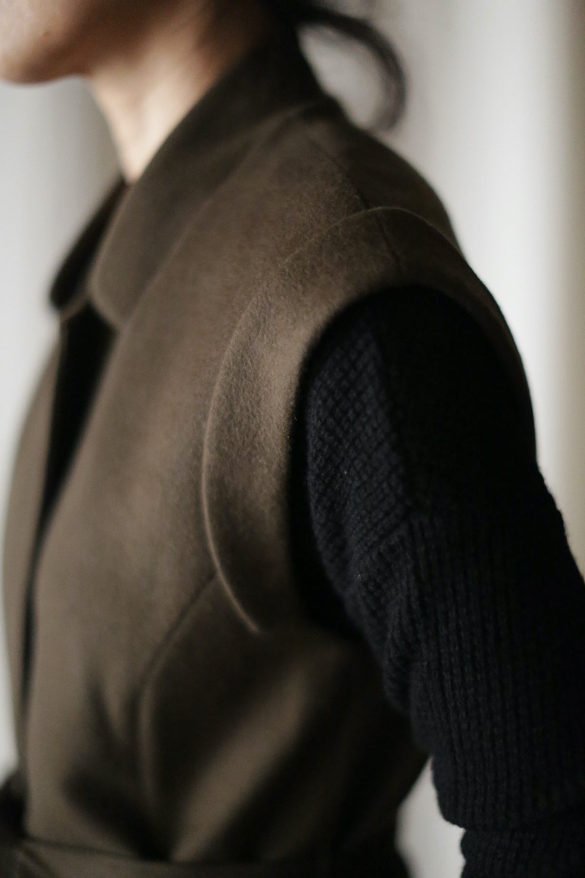 A close-up of a person wearing the Lexington, a dark olive-brown sleeveless coat with a belted waistline over a black long-sleeve knit top, focusing on the shoulder and upper arm against a softly blurred background.