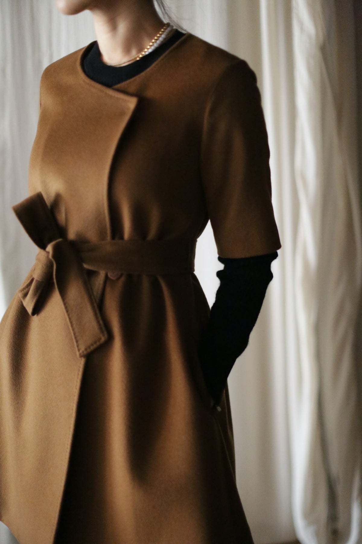 A person in a stylish, brown Wexley cashmere coat with an A-line cut and short sleeves over a black top. The minimalist design is enhanced by a belt, set against soft, flowing white curtains.