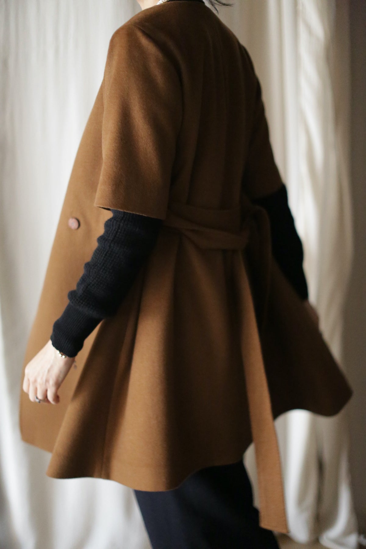 A person stands in front of light curtains, wearing the Wexley—a brown, knee-length double-breasted woven cashmere coat with a back-tied belt—over a black long-sleeve top.