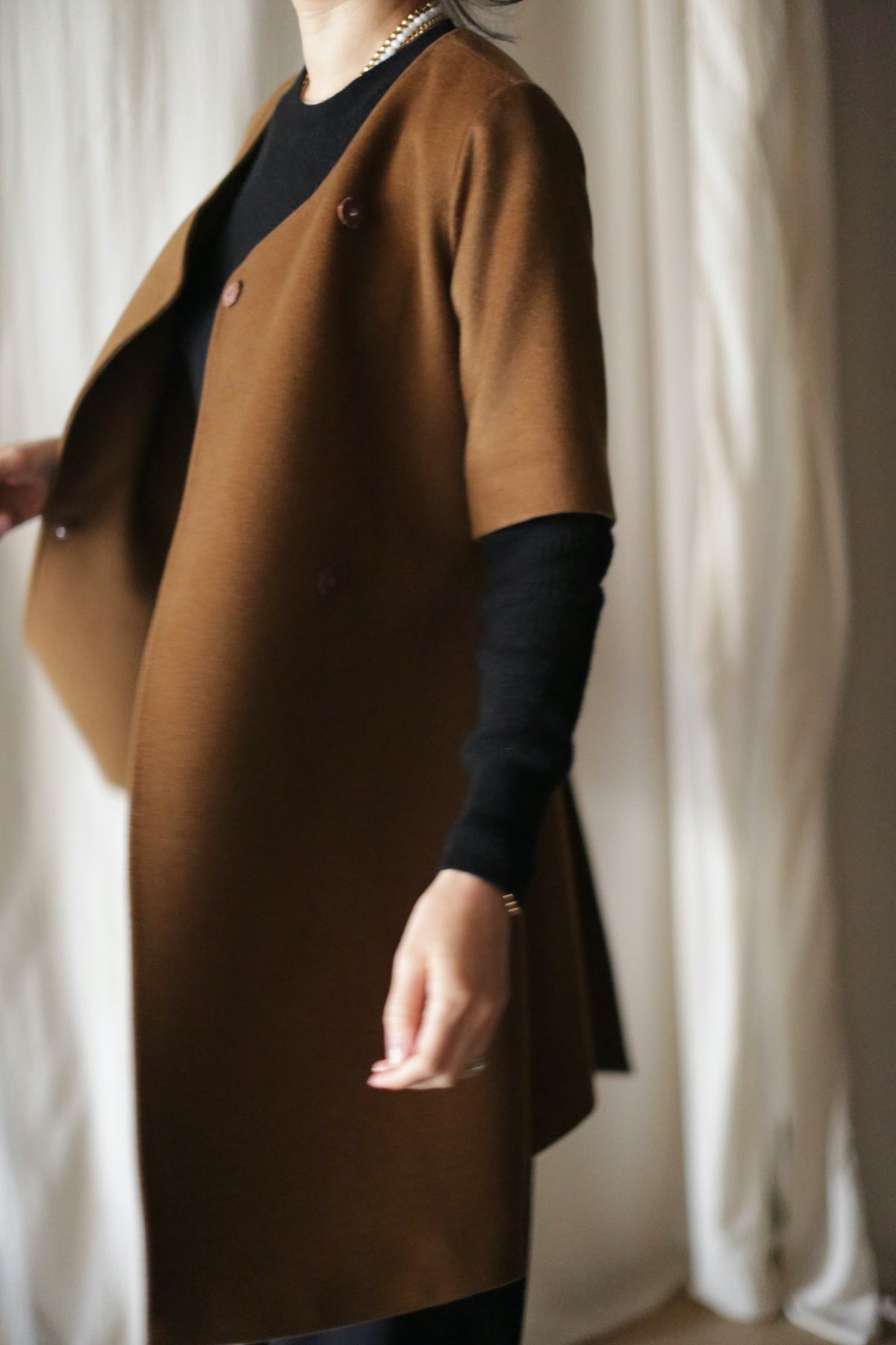 A person stands in a softly lit room with light-colored drapes, wearing the stylish Wexley brown double-breasted coat over a black long-sleeve top. Their face is not visible.