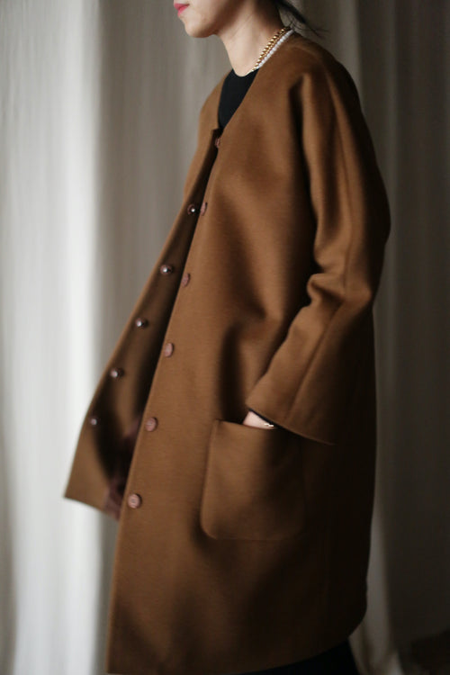A person stands sideways wearing the Battersea long brown cashmere coat with raglan sleeves and large handmade snap buttons, their left hand in a pocket. The neutral-toned background is softly blurred.