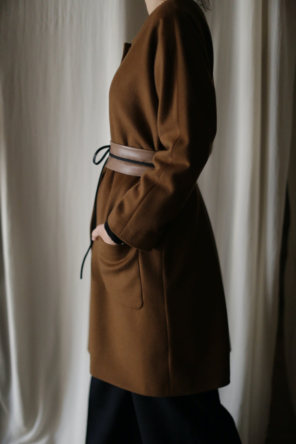 A person stands in profile against a light, softly textured background, wearing the Battersea brown cashmere coat with raglan sleeves and a belted waist; only the side is visible, with one hand in the pocket.
