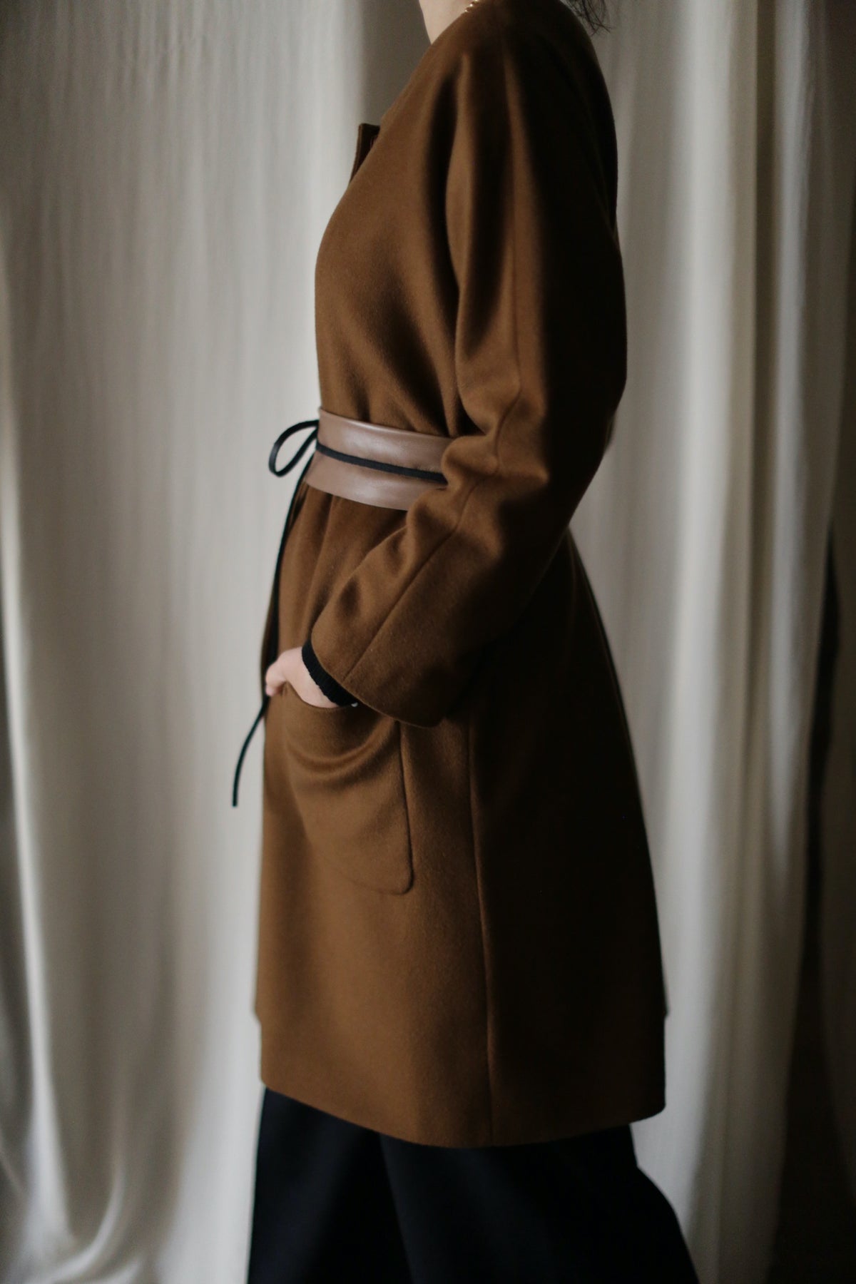 A person stands in profile against a light, softly textured background, wearing the Battersea brown cashmere coat with raglan sleeves and a belted waist; only the side is visible, with one hand in the pocket.