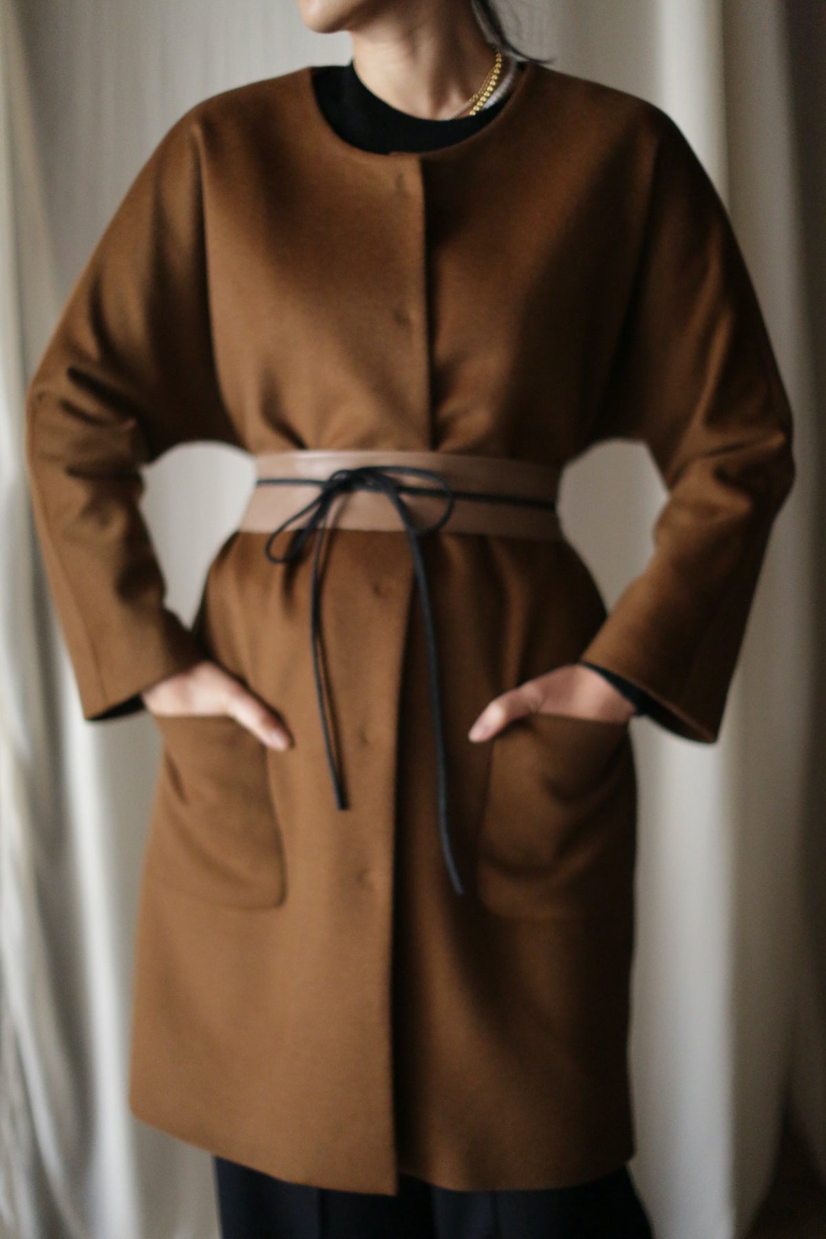 Someone stands against a light-colored curtain background, wearing the Battersea—a brown cashmere coat with large front pockets and raglan sleeves, cinched at the waist with a thin beige belt tied with a black string.