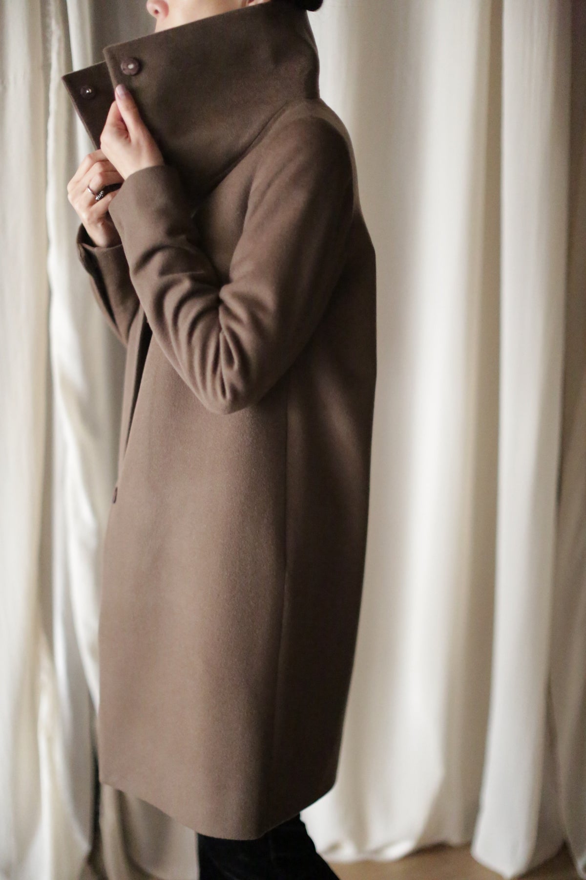 A person wearing the Stamford Long cashmere coat stands with their side facing the camera, gently holding the collar up with one hand. Light curtains create a soft backdrop.