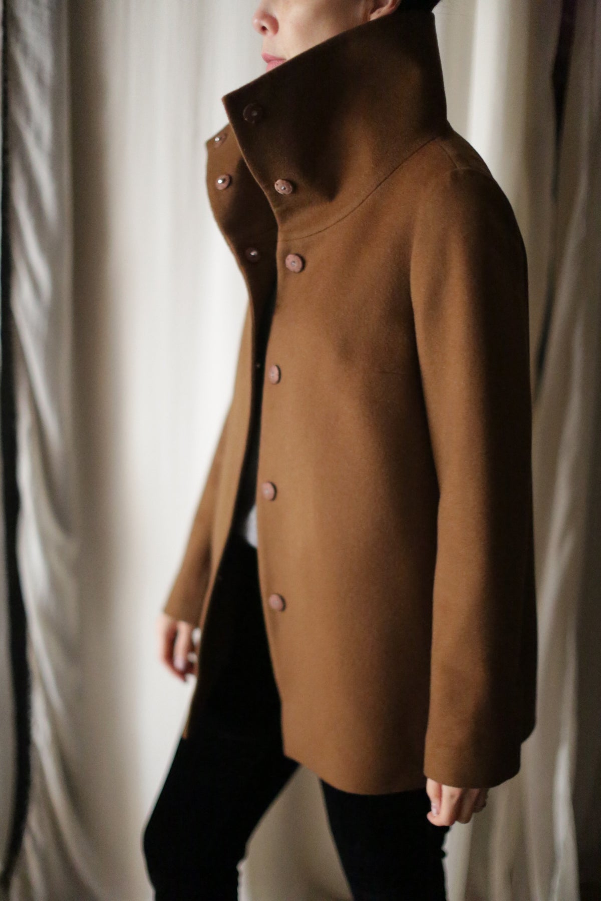 A person wearing the Stamford II jacket stands in profile against a neutral background. The high-collared, brown button-up coat is crafted from woven cashmere and features a minimalist design with simple buttons. The individual appears to be indoors, with only their torso and part of their face visible.