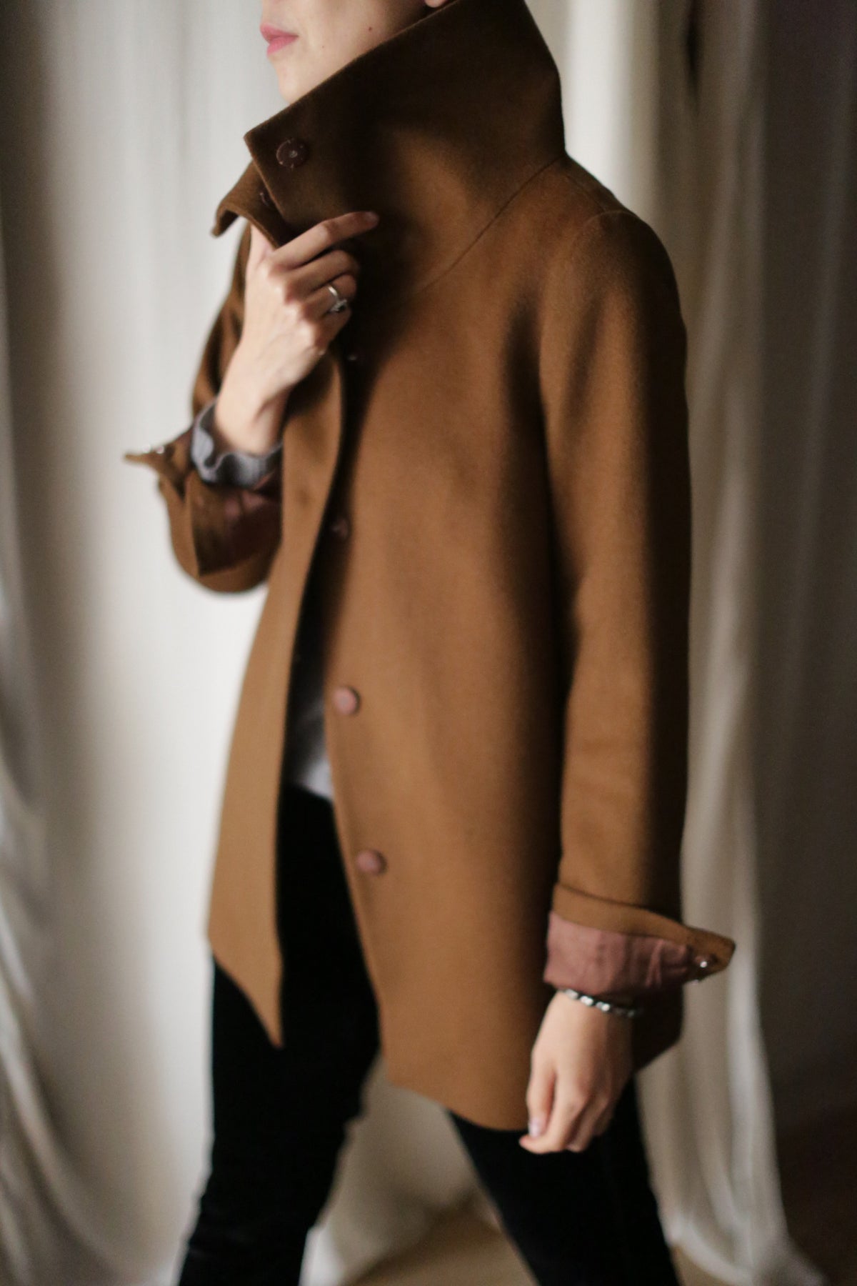 A person is standing indoors, partially turned away from the camera. They are wearing a Stamford II jacket in brown with the high collar turned up, covering part of their face. The individual is gripping the collar with one hand while their other arm is bent at the elbow.