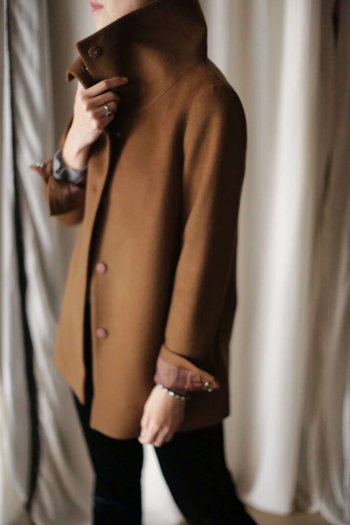 An individual indoors, captured in side profile, wears a Stamford II jacket with its high woven cashmere collar turned up. Their hand gently touches the collar while white and beige curtains create a blurred background.
