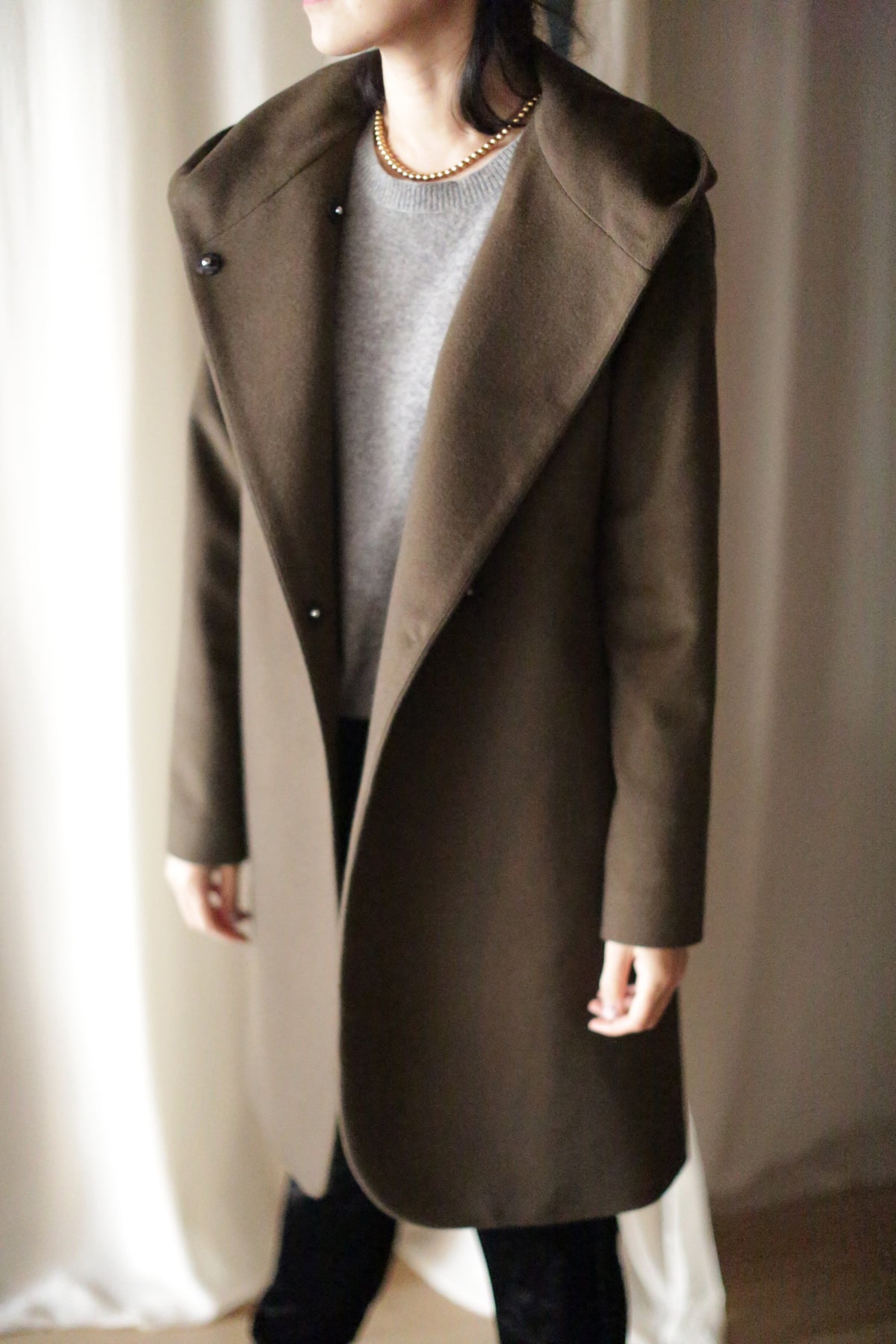 A person gracefully stands before a light curtain, wearing the Mitchley Long coat, a stylish brown piece with fairytale-like length over a gray sweater. It features a wide collar and dark handmade snap buttons, pairing elegantly with their dark hair.
