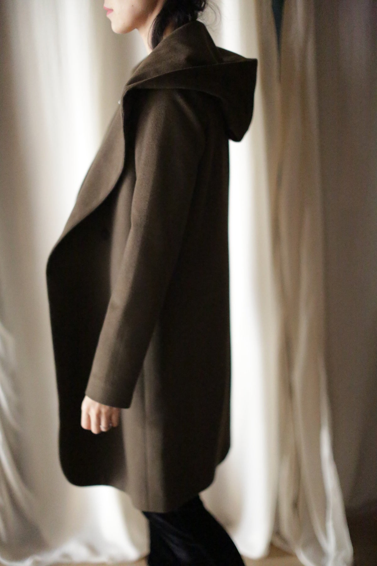 Wearing the Mitchley Long dark brown coat with a hood and handmade snap buttons, a person stands in profile against white curtains. Their face is partially visible, long dark hair falls freely, and the soft lighting creates a warm, fairytale-like shadow.