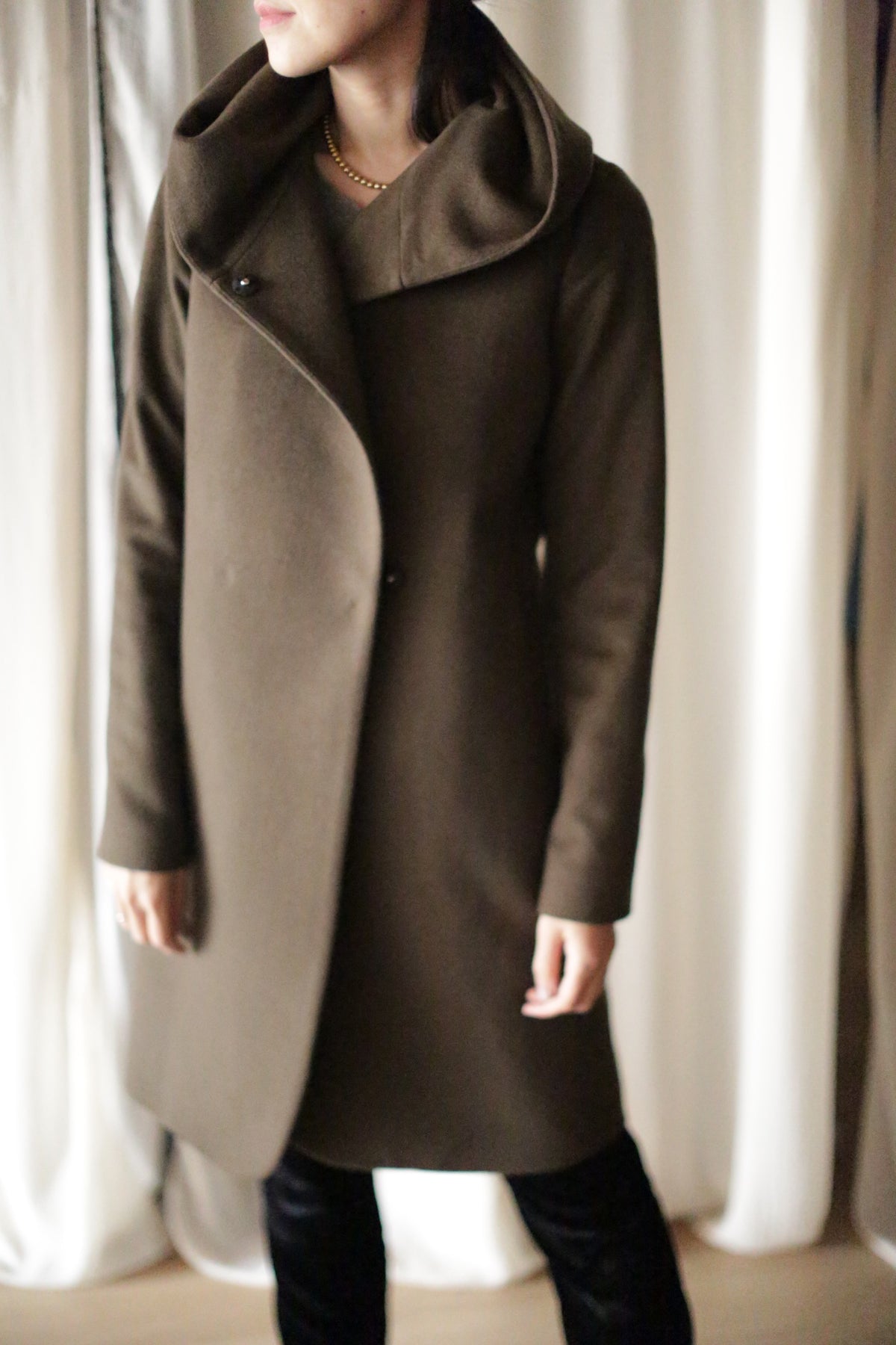 Wearing the Mitchley Long, a stylish dark olive green coat with an asymmetrical front, hood, and handmade snap buttons, the person stands against cream-white curtains. The fairytale-like length enhances its allure while their face remains unseen.