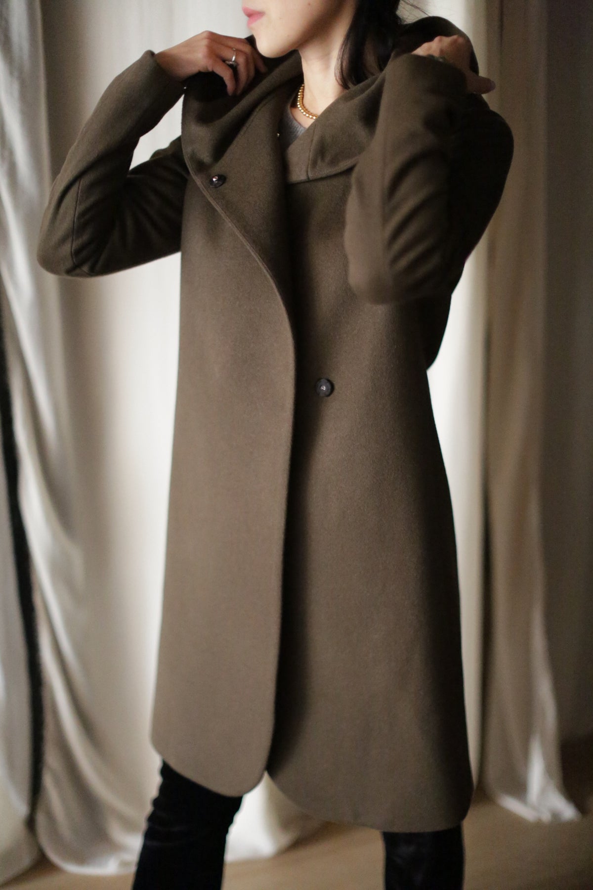 A person adjusts the collar of their Mitchley Long olive-green cashmere coat, featuring handmade snap buttons. It's buttoned up against a backdrop of draped white curtains, with their face obscured.