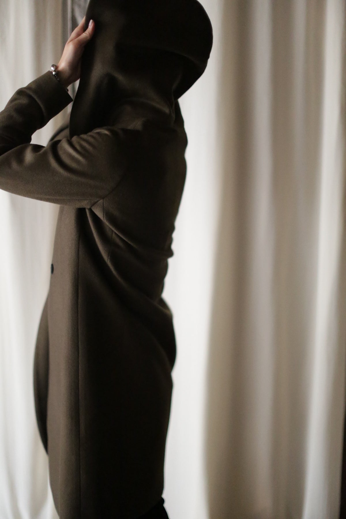 A person wearing the Mitchley Long coat with handmade snap buttons is partially facing away, pulling the hood over their head. Light-colored curtains form the backdrop, creating a subdued mood focused on the act of putting on the hood.