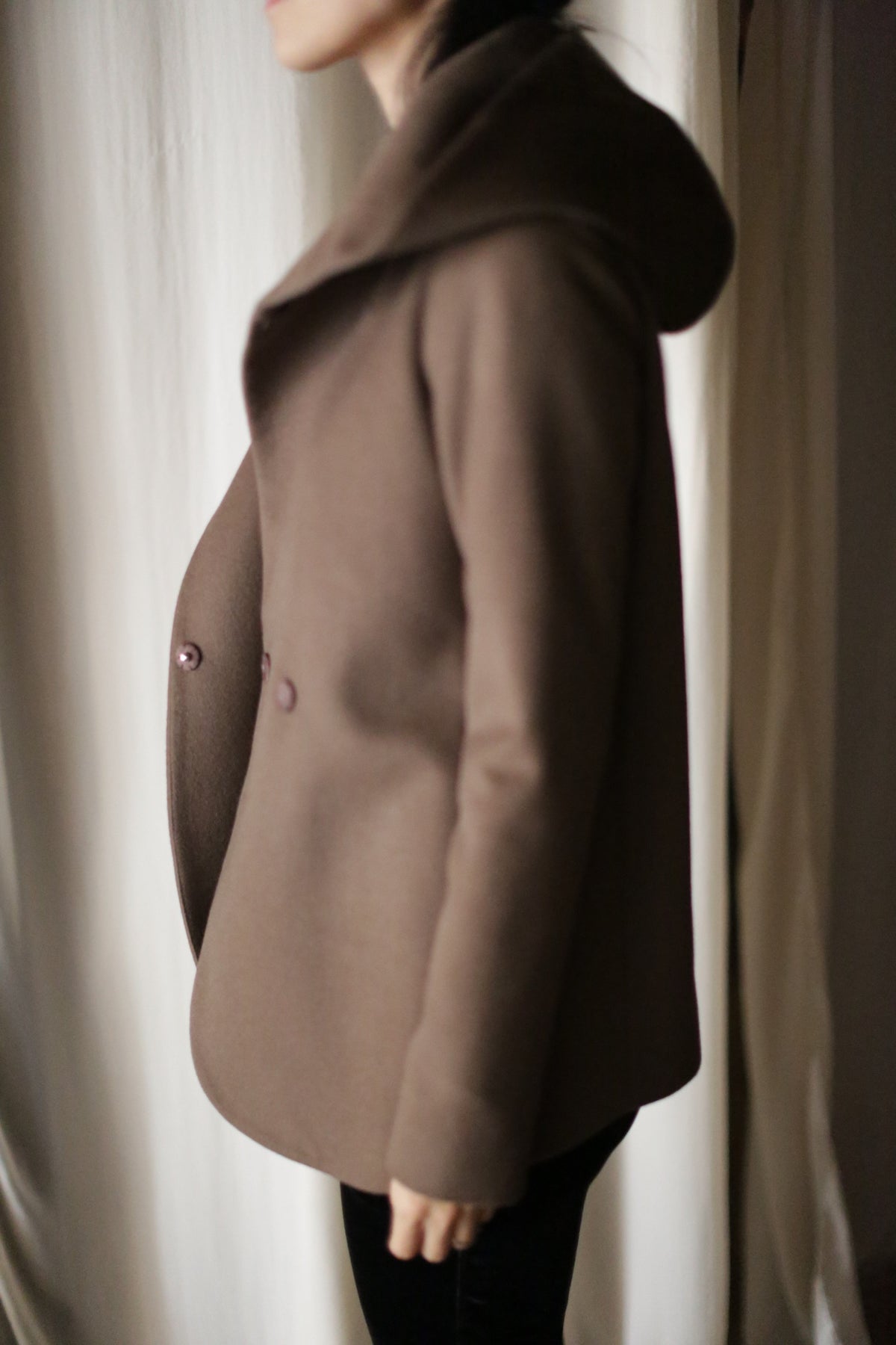 A person stands sideways in the Mitchley—a luxurious brown, double-breasted jacket with large lapels and a cashmere hood—against a light curtain. Their face is partially out of frame.