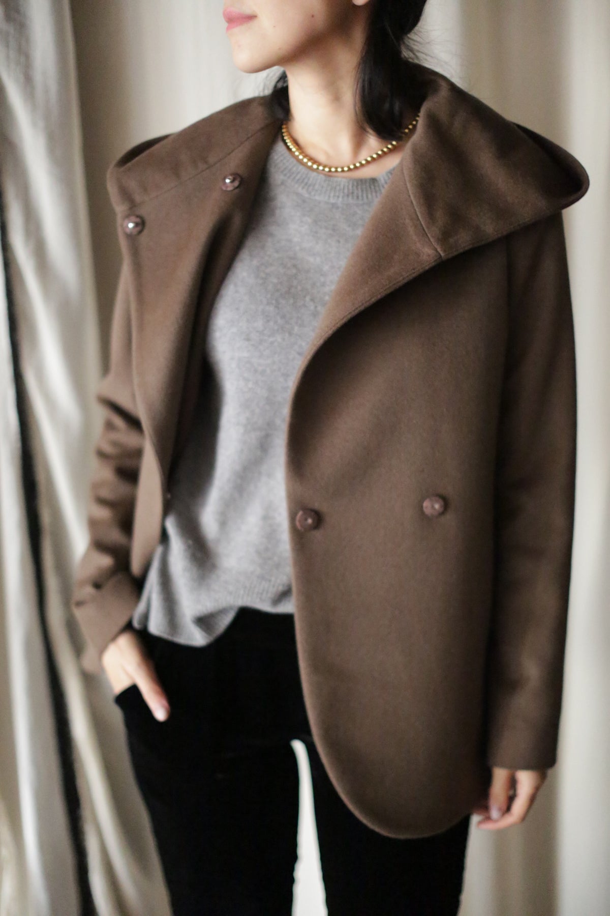A woman wears the Mitchley jacket—a brown, double-breasted design with a wide collar—over a gray sweater and black pants, accessorized with a gold necklace. She stands with one hand in her pocket; her face is partially visible.