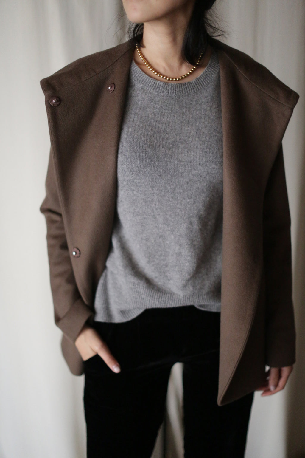 A person stands against a light background, wearing the luxurious Mitchley jacket over a gray sweater, black pants, and a gold beaded necklace. Their hand is in one pocket and their face is partially out of frame.