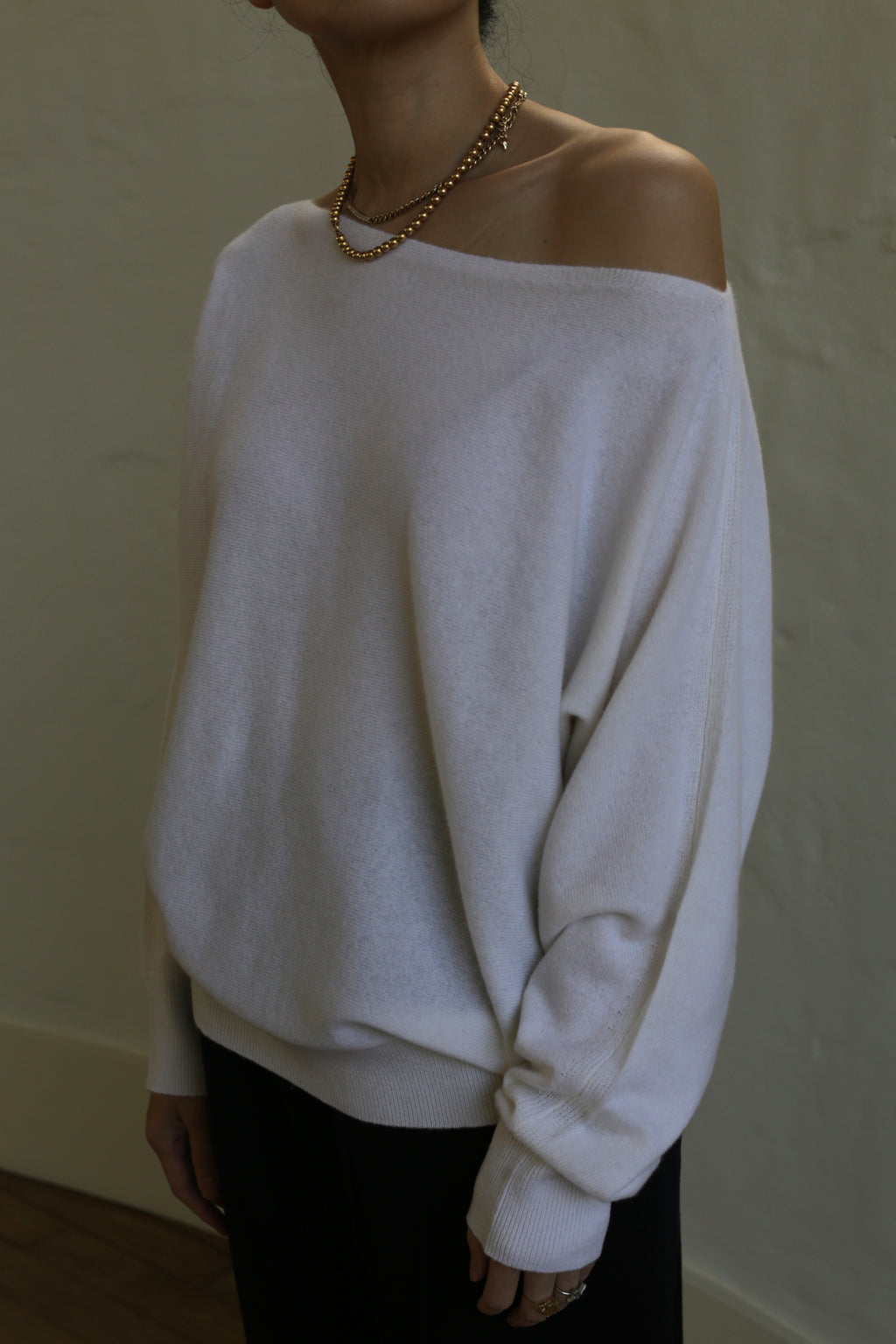 A person wearing an off-the-shoulder, relaxed-fit, Ballerina's Cashmere Sweater | Ivory paired with black pants. They are accessorized with a gold chain necklace and several rings. The background is a soft, neutral-colored wall. The person's head is not visible in the image.